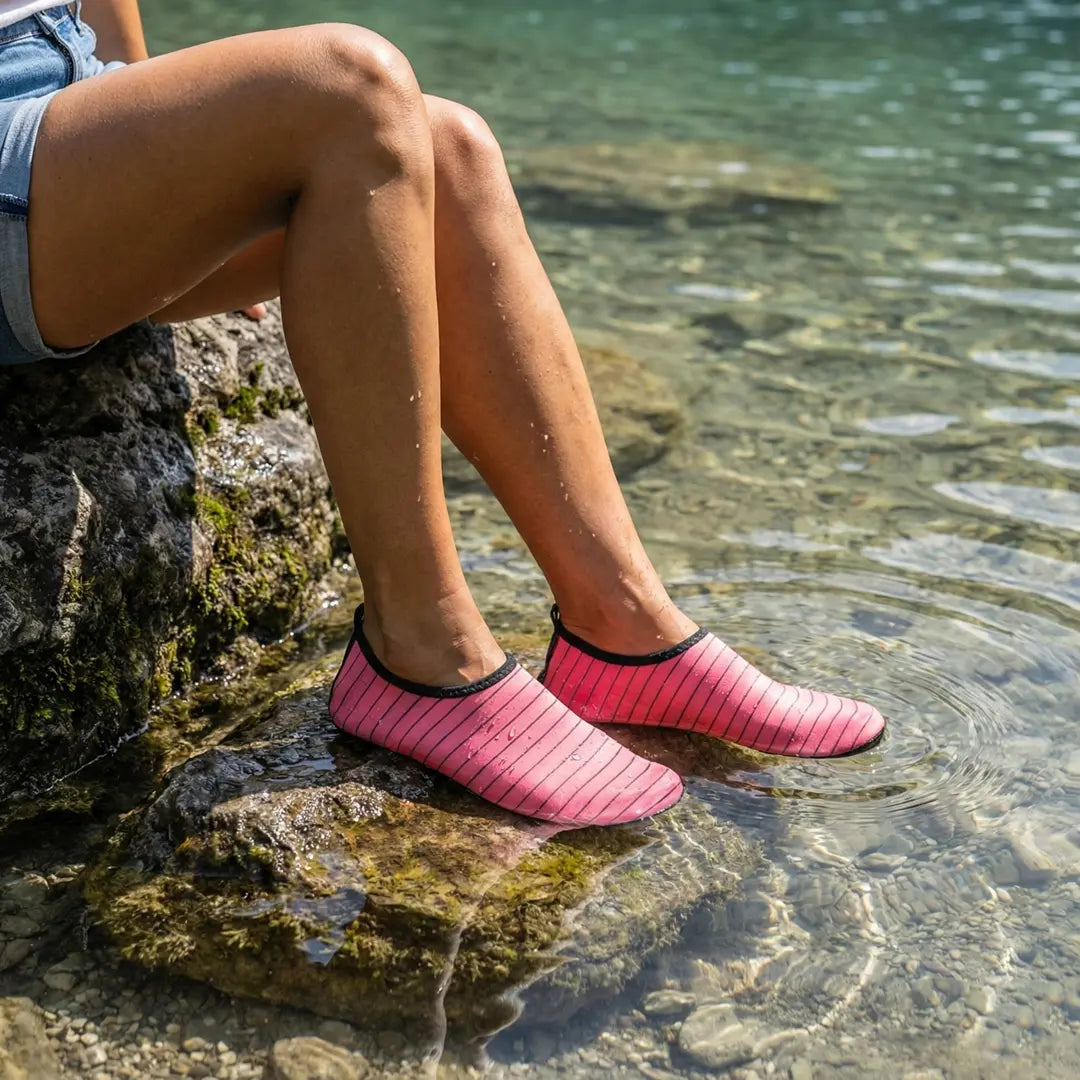Mujer sentada en una roca con los pies en el agua usando zapatillas rosas.