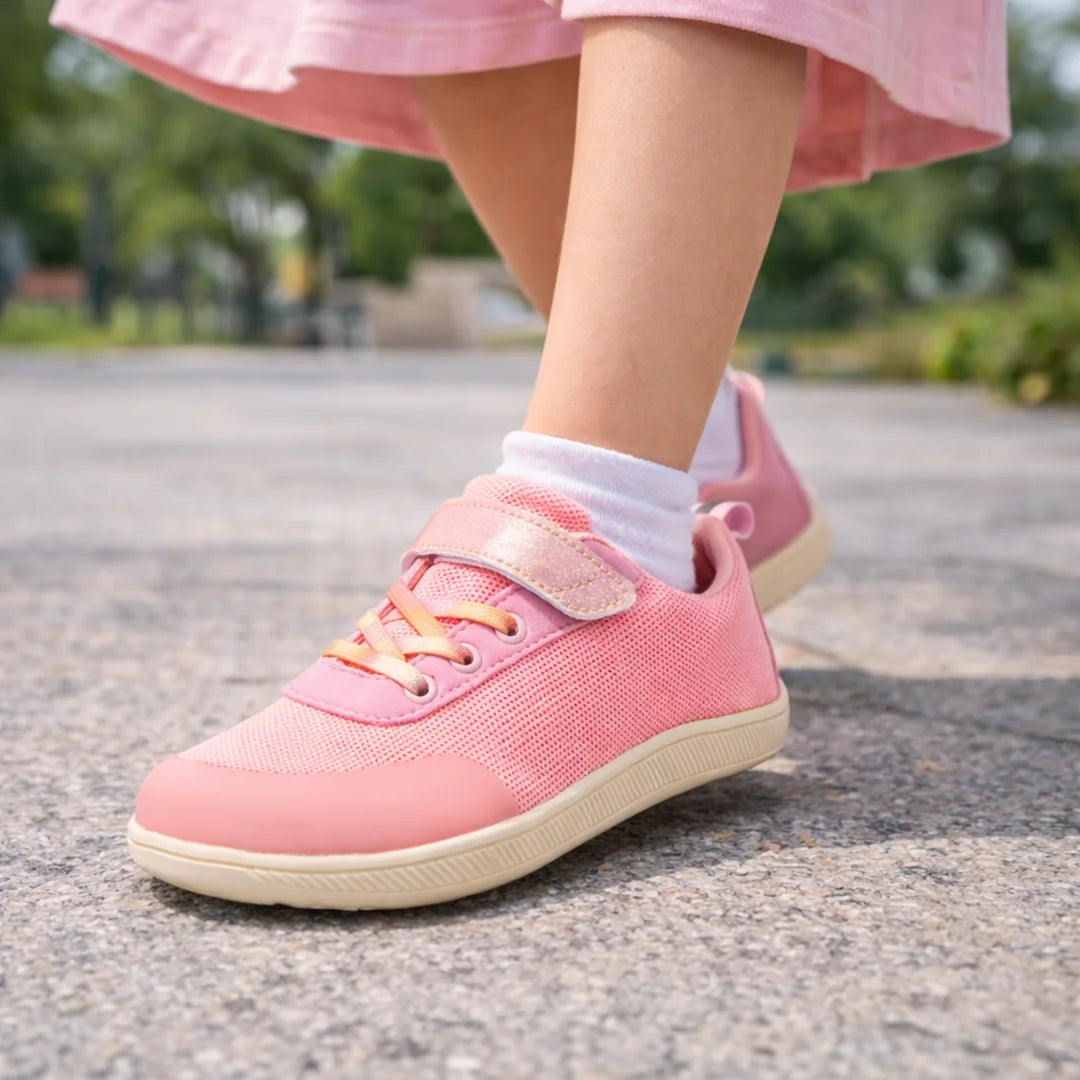 Vista de perfil de calzado barefoot rosa para niña en un entorno de parque y juego.